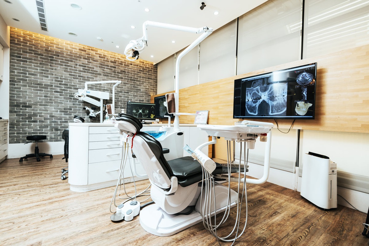 A dental chair sits at the center of a dentist's office, surrounded by a desk and a few computer screens.