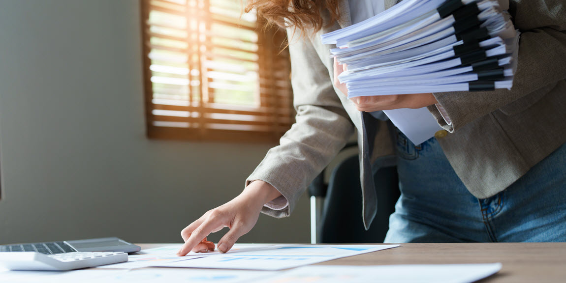 Accountant standing at desk with large stack of papers in one arm.  